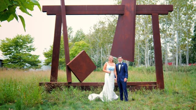 A couple in wedding attire poses in front of a large outdoor metal sculpture surrounded by greenery.