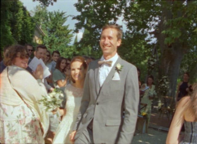 A happy bride and groom walking down the aisle at an outdoor wedding, surrounded by smiling guests. The groom wears a grey suit and the bride a lace dress, both under a clear blue sky.