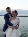 Bride and groom embracing by the water at sunset, with the bride smiling as the groom holds her from behind.